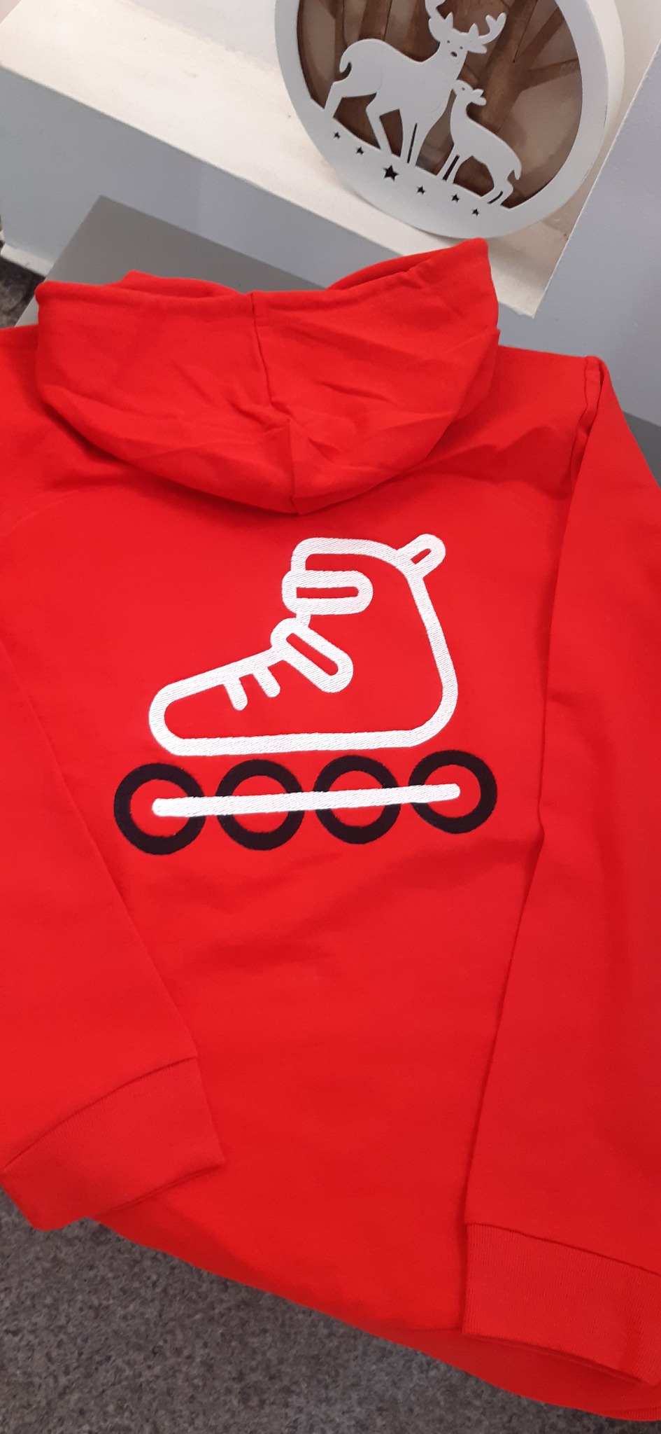 Czerwona bluza z kapturem, na plecach biały nadruk przedstawiający rolkę. U góry dekoracja świąteczna z motywem reniferów.