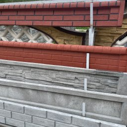 Mono-Steel - Elementy betonowe ogrodzenia imitujące cegłę i kamień, składowane jeden na drugim. Widoczne różne wzory i kolory, przygotowane do montażu. Tło: zieleń i fragment dachu.