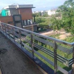 Mono-Steel - Nowoczesna balustrada nierdzewna z poziomymi elementami, zamontowana na balkonie z widokiem na zielony ogród i dom w tle. Metaliczna powierzchnia odbija światło.