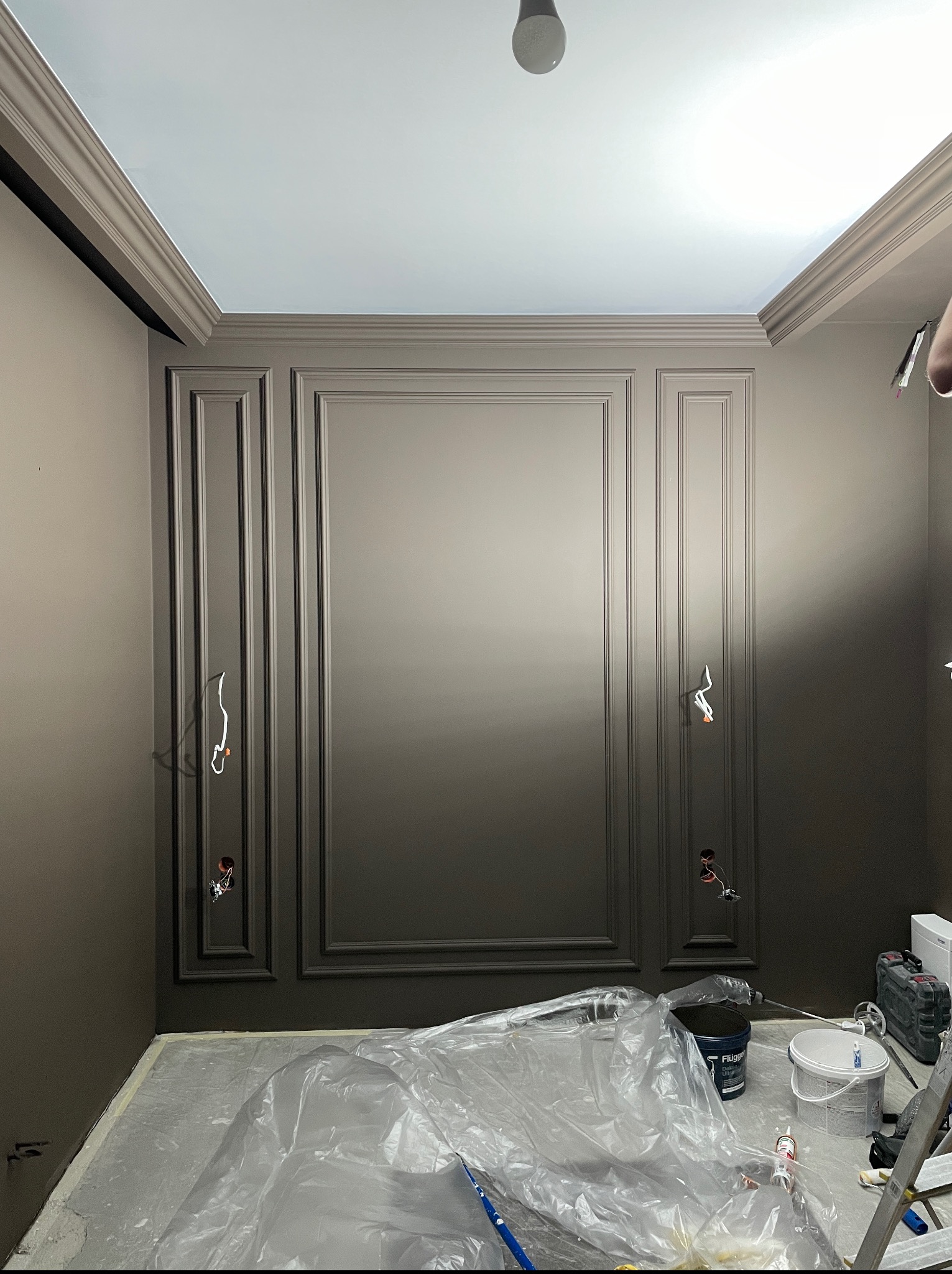 Ściana z dekoracyjnymi panelami pomalowana na jednolity, ciemny odcień brązu. Podłoga zabezpieczona folią malarską. Widoczne puszki elektryczne bez osprzętu.