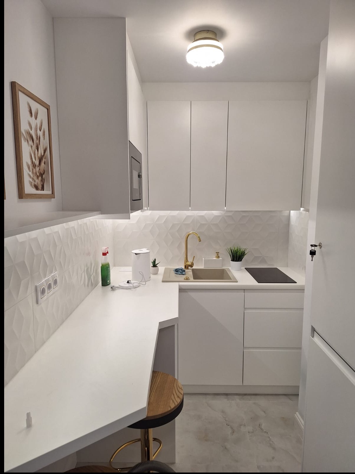 Biała, nowoczesna kuchnia z geometrycznymi płytkami, złotą armaturą i drewnianym stołkiem barowym. Minimalistyczny design z oświetleniem LED i roślinami doniczkowymi.