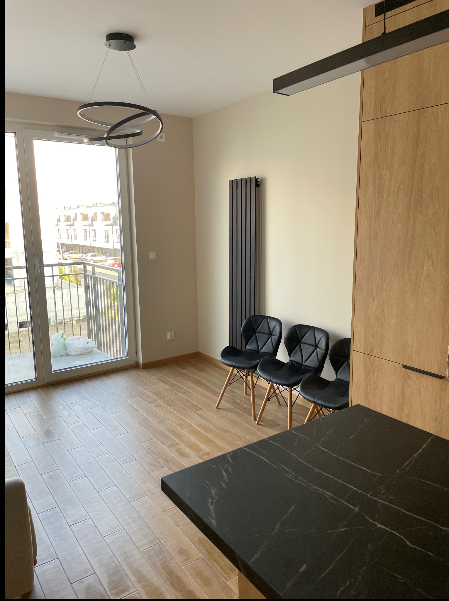 Jasne wnętrze z drewnianą podłogą, trzema czarnymi krzesłami i nowoczesną lampą. Widok na balkon i zabudowę kuchenną z blatem imitującym czarny marmur.