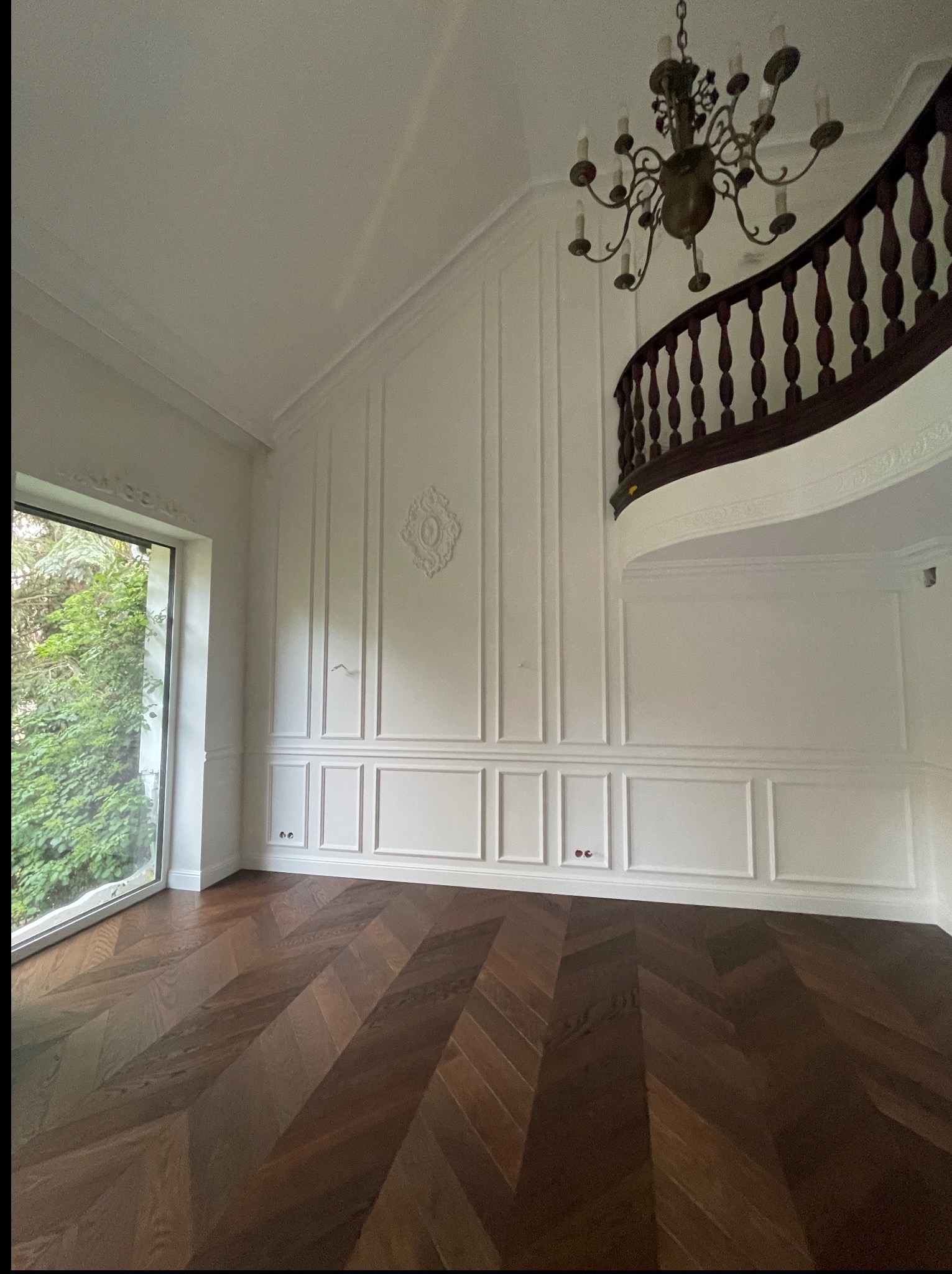 Eleganckie wnętrze po remoncie: parkiet w jodełkę, białe ściany z dekoracyjnymi panelami, widok na schody z drewnianą balustradą i żyrandol.