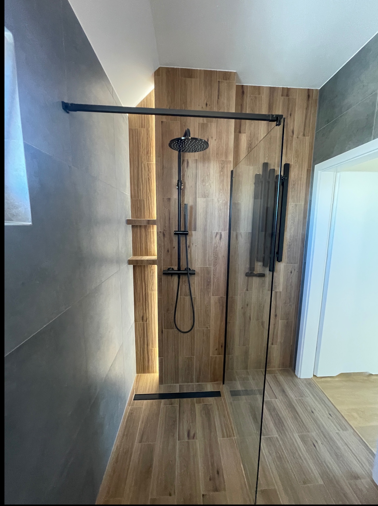 Nowoczesna kabina prysznicowa walk-in z czarną armaturą i drewnopodobnymi płytkami, oświetlona nastrojowym światłem. Minimalistyczny design z dbałością o detale.