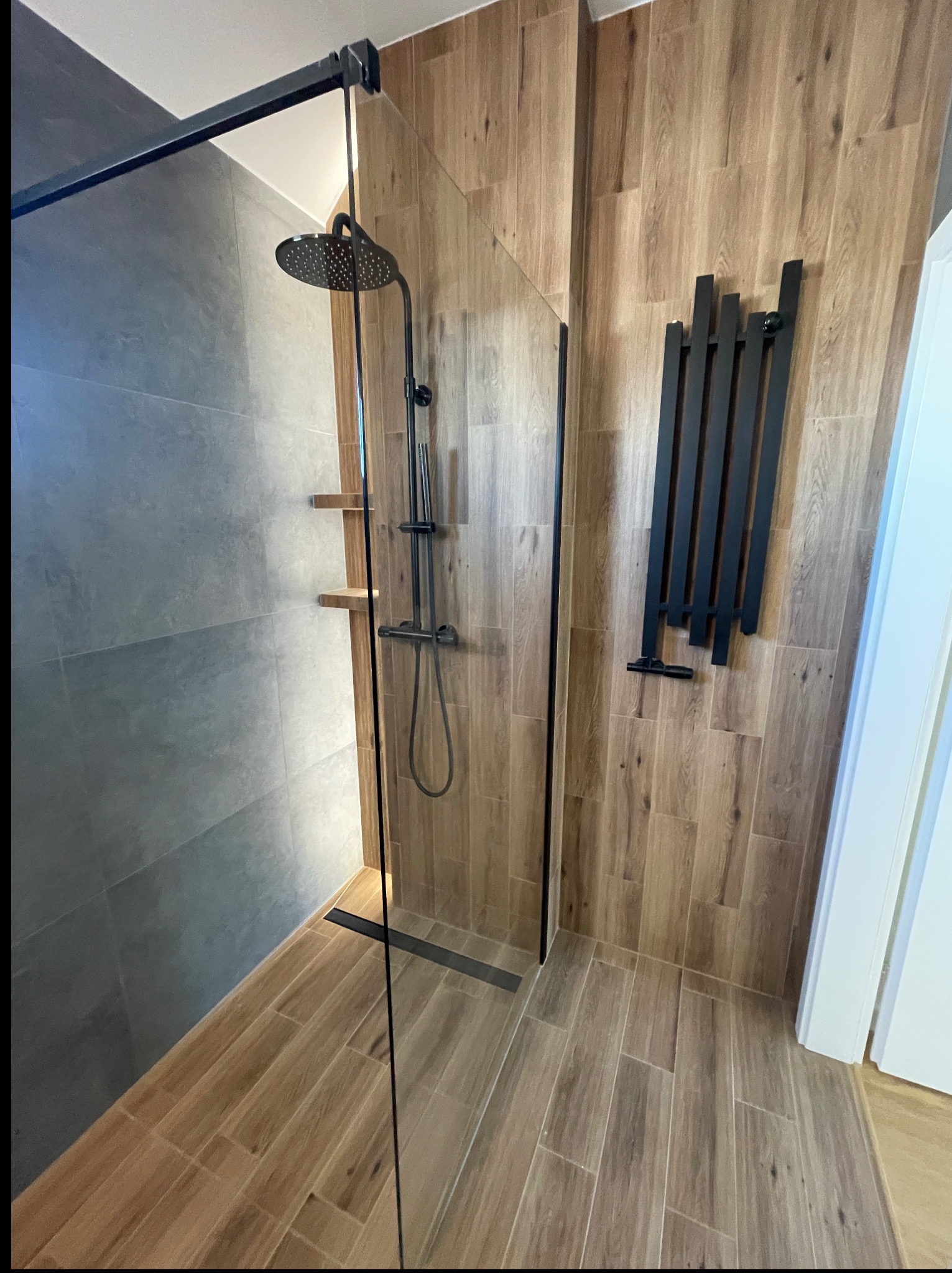 Nowoczesna kabina prysznicowa typu walk-in z czarnymi elementami i drewnopodobnymi płytkami na ścianach, uzupełniona o grzejnik drabinkowy.