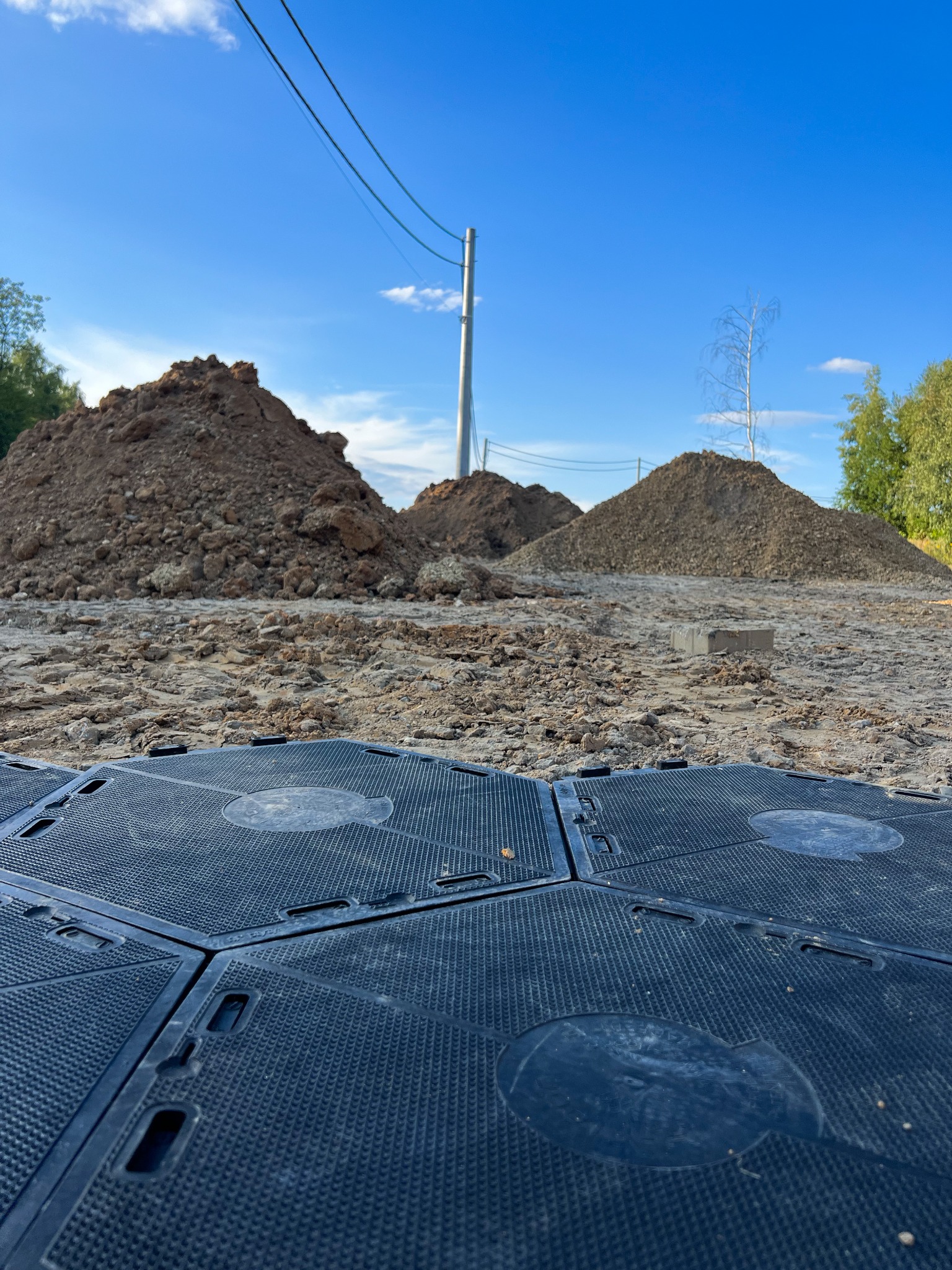 Tymczasowa nawierzchnia z czarnych, modułowych płyt na piasku, z pryzmami ziemi w tle pod błękitnym niebem. Teren budowy lub imprezy plenerowej.