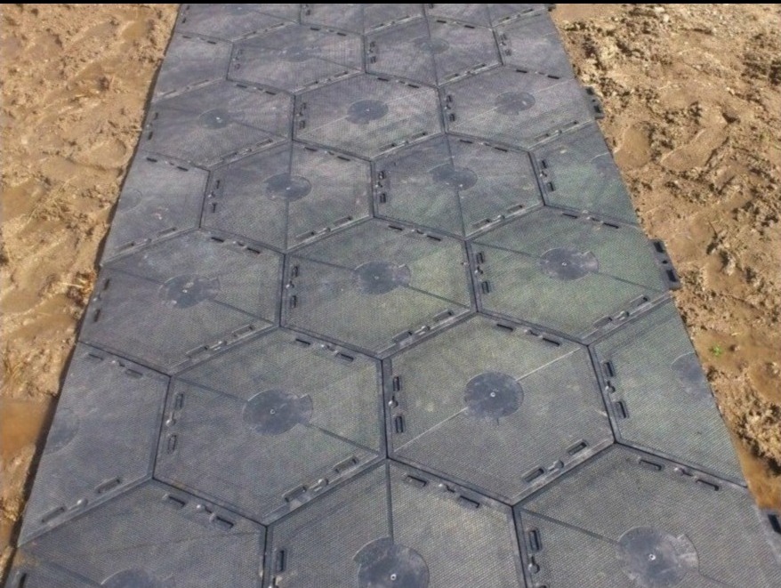 Ułożona, tymczasowa podłoga z heksagonalnych paneli na grząskim terenie. Panele w kolorze ciemnoszarym, połączone ze sobą, tworzą ścieżkę.