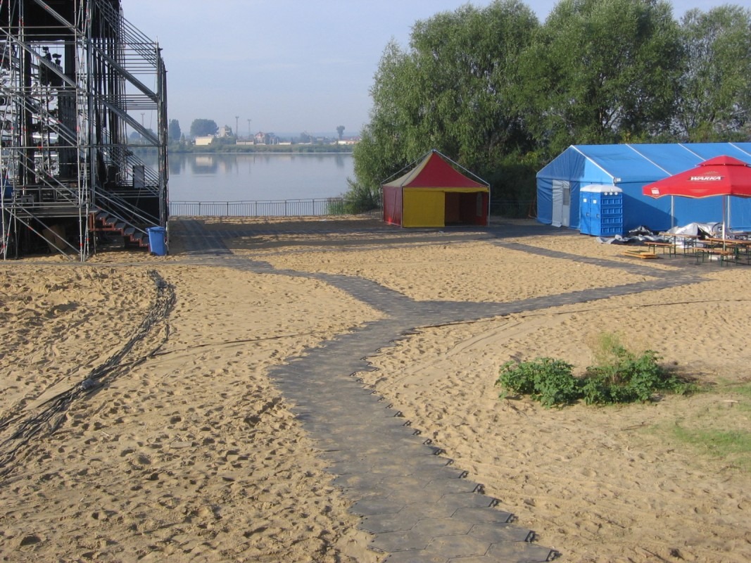 Piaszczysta plaża z przygotowanym terenem pod imprezę plenerową: scena, namioty, przenośna toaleta i podłoże z paneli. W tle jezioro.