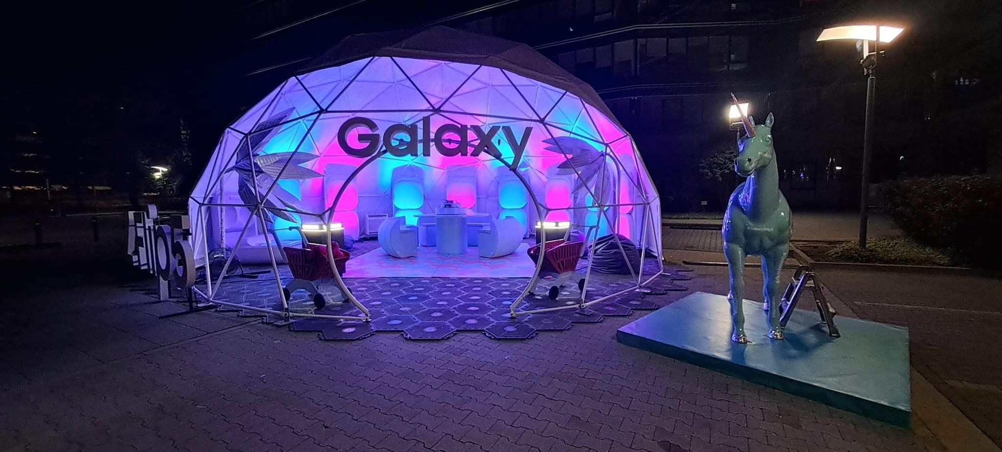 Iluminowany geodezyjny namiot eventowy z logo Galaxy, ustawiony na zewnątrz, obok rzeźby konia, podświetlony lampą uliczną. Nocne ujęcie.