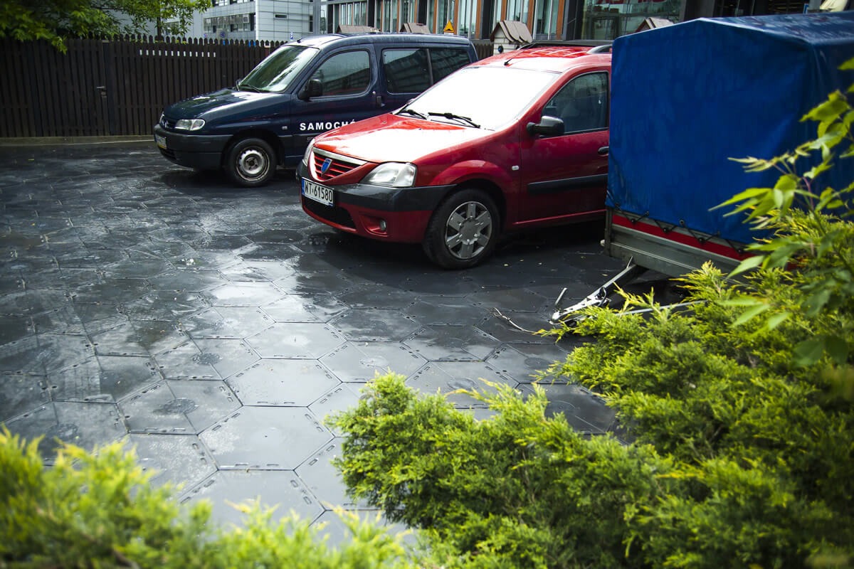 Ułożona, sześciokątna nawierzchnia z szarego tworzywa, częściowo mokra, z zaparkowanymi samochodami w tle. Widok zza krzewów.
