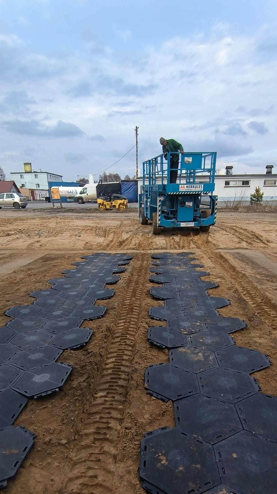 Montaż tymczasowej nawierzchni z heksagonalnych płyt na gruncie. Pracownik na podnośniku koszowym. W tle budynki i maszyny budowlane.
