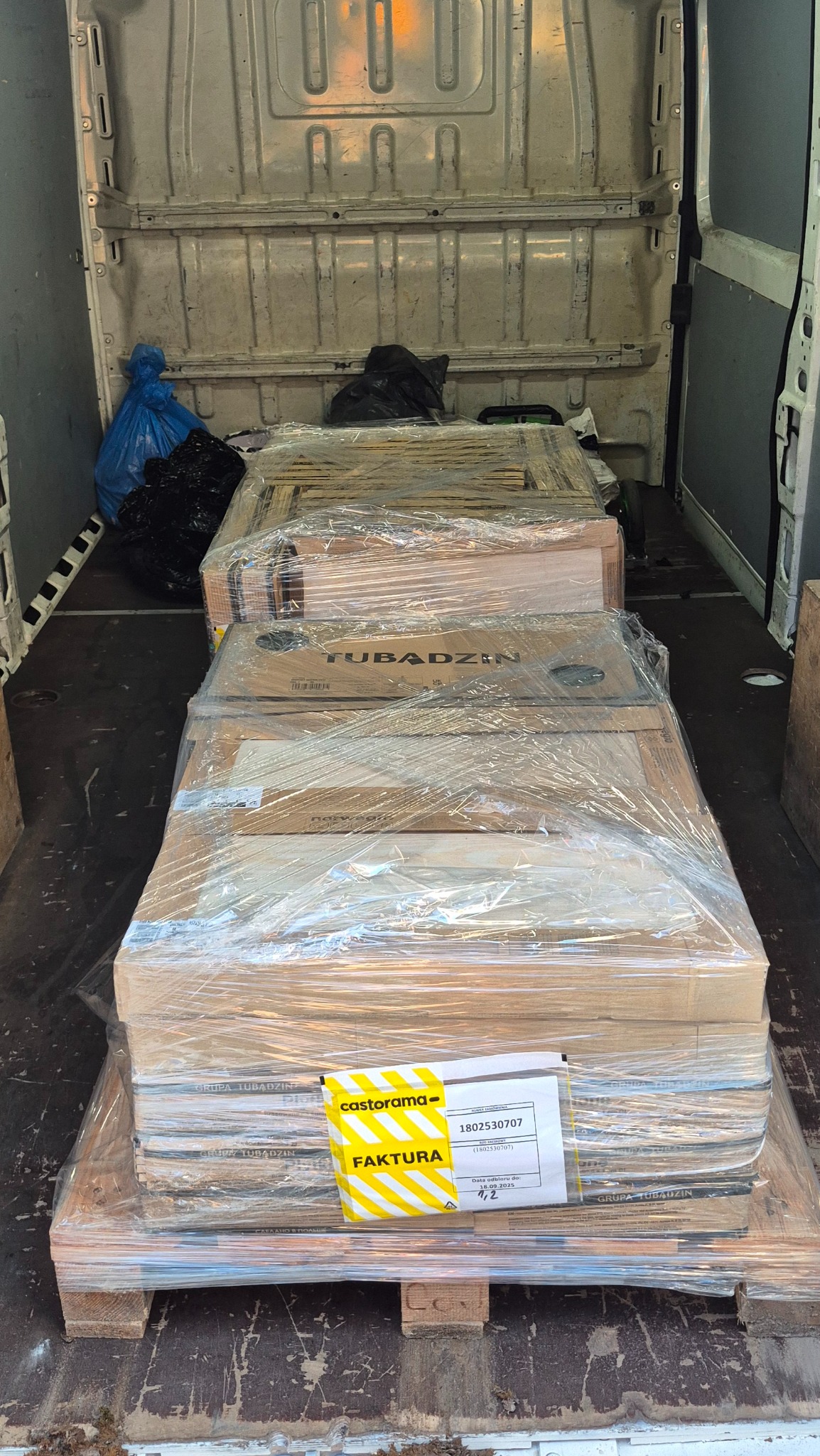 Palety z zapakowanymi płytkami Tubądzin w przestrzeni ładunkowej busa, zabezpieczone folią stretch i opatrzone fakturą z Castoramy. Widoczne worki w tle.