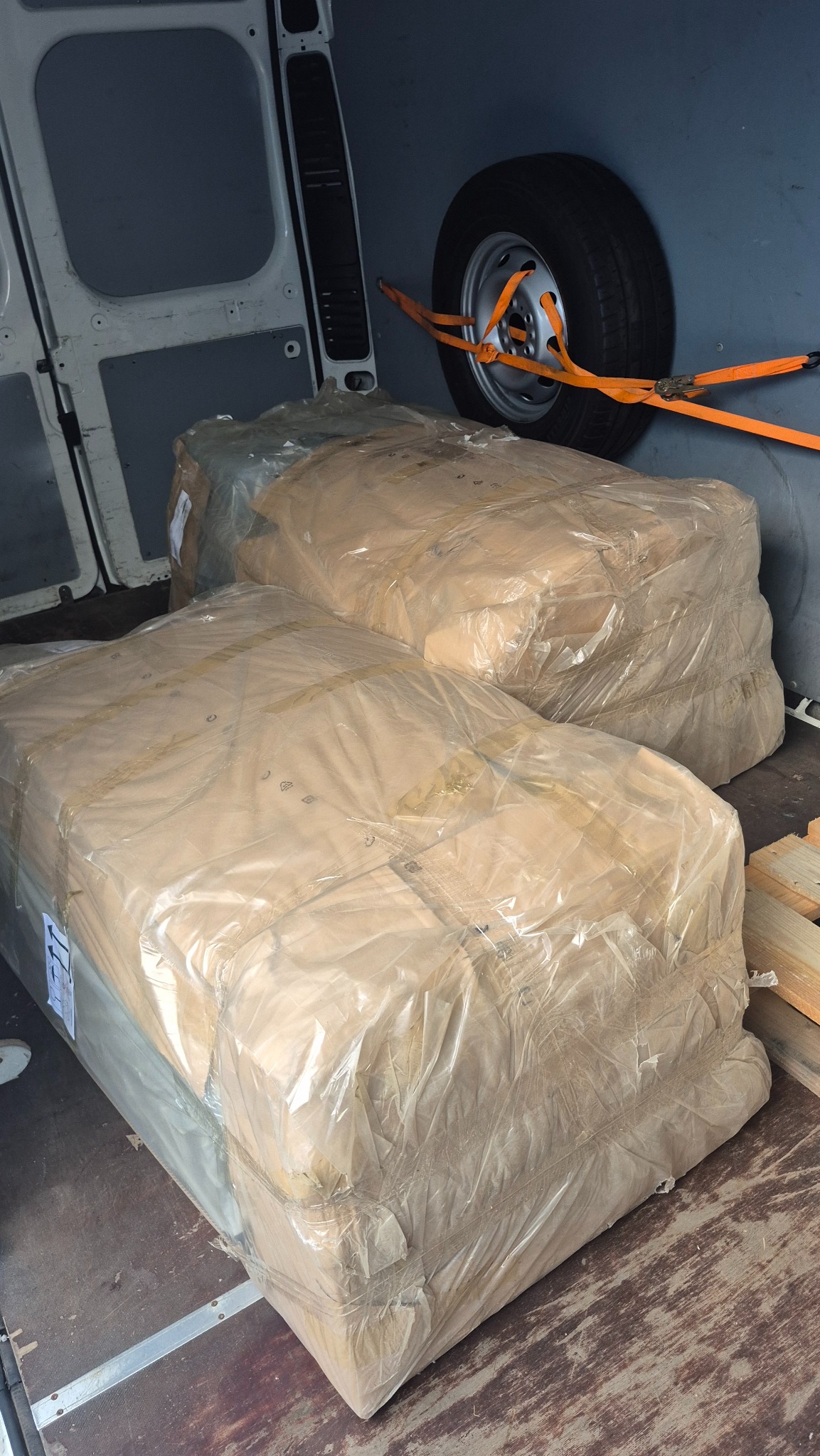 Dwa zapakowane, zabezpieczone paczki wewnątrz pojazdu transportowego, z kołem zapasowym przymocowanym do ściany za pomocą pomarańczowych pasów.