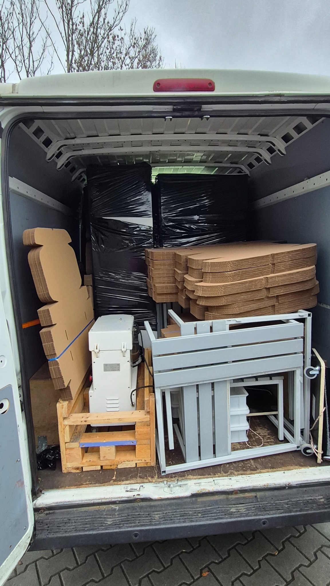 Wnętrze busa załadowane kartonami, meblami w metalowych ramach i sprzętem owiniętym folią, gotowe do transportu. Widok z tyłu pojazdu.