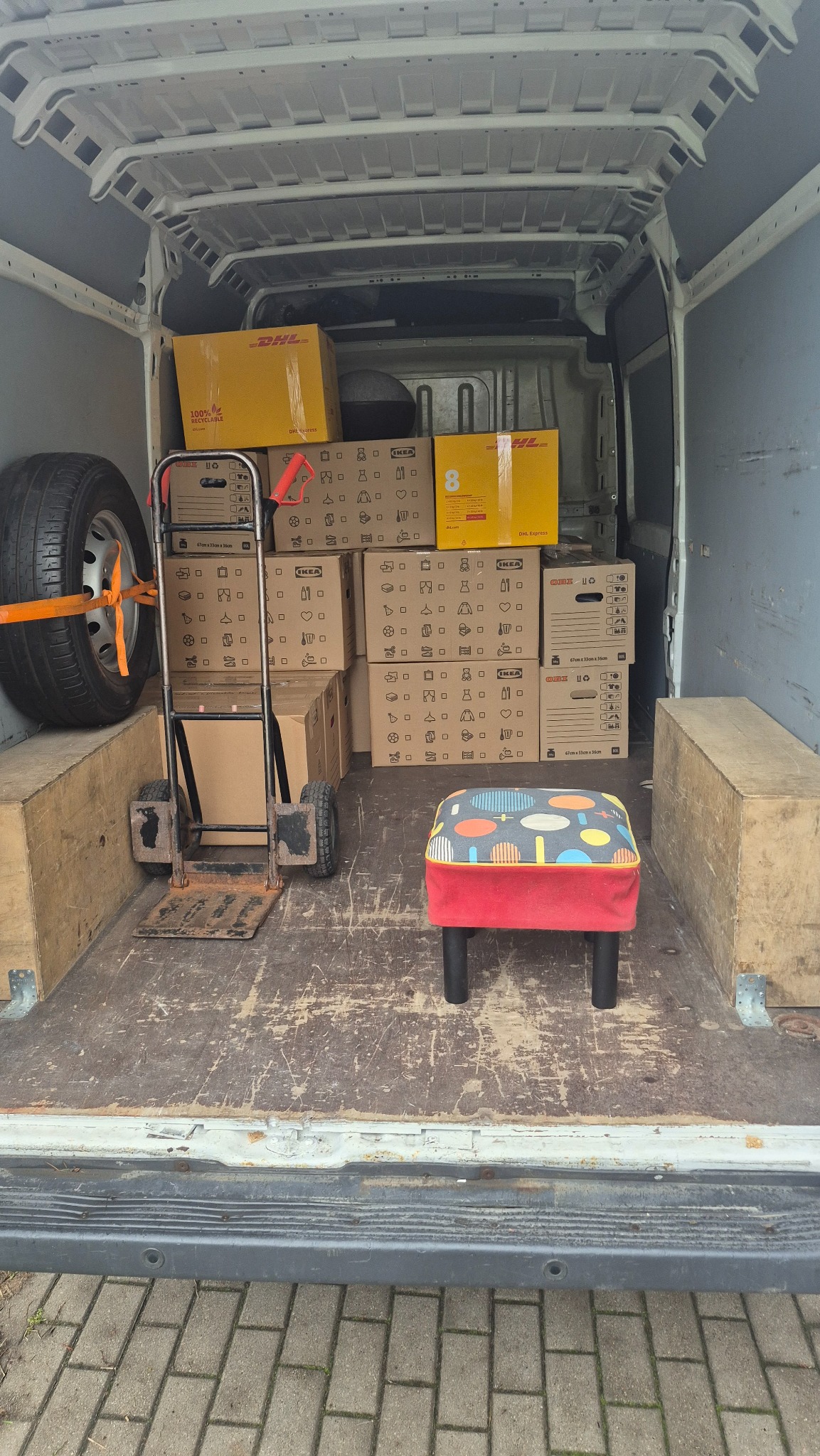 Wnętrze busa załadowane kartonami DHL i IKEA, wózek transportowy i pufa. Koło zapasowe zabezpieczone pasem. Widok z tyłu pojazdu.