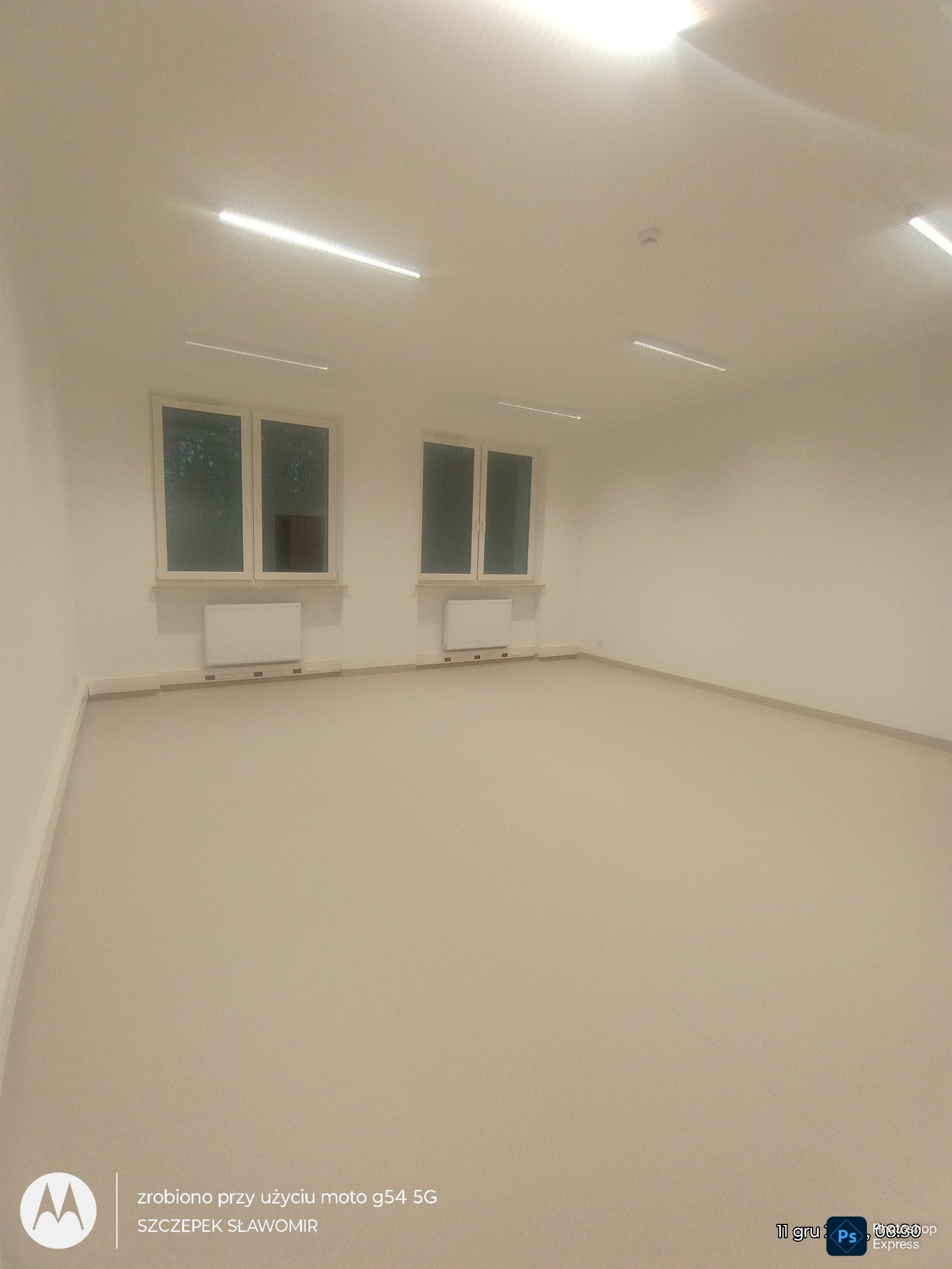 Wnętrze po remoncie: puste, białe pomieszczenie z dwoma oknami, grzejnikami i jasną podłogą. Widoczne listwy przypodłogowe i oświetlenie sufitowe.