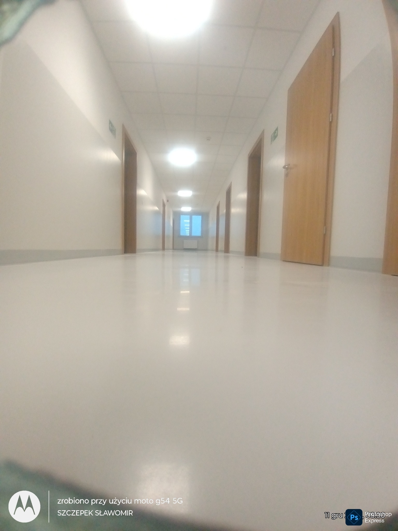 Długi korytarz z jasnymi ścianami i podłogą, rzędy drewnianych drzwi po obu stronach, oświetlony sufit z równomiernie rozmieszczonymi lampami. Perspektywa z niskiego punktu widzenia.