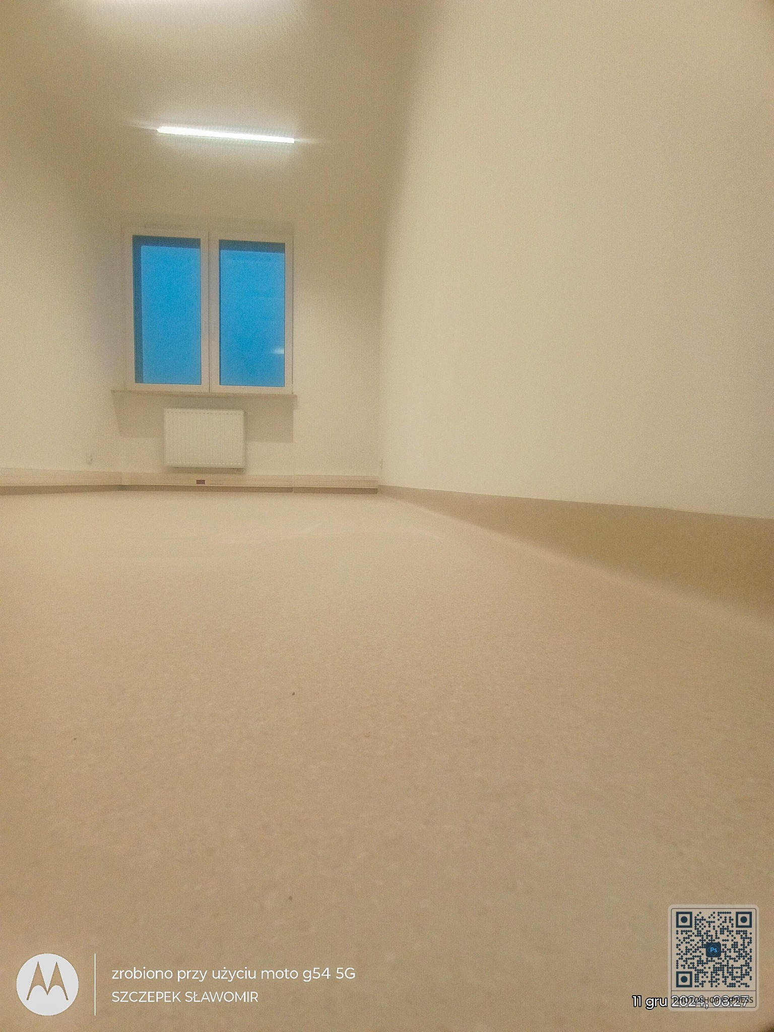 Pomalowane na beżowo wnętrze pokoju z oknem i grzejnikiem. Widoczna listwa przypodłogowa. Świeżo wykończone ściany i podłoga. Całość oświetlona światłem z lampy sufitowej.