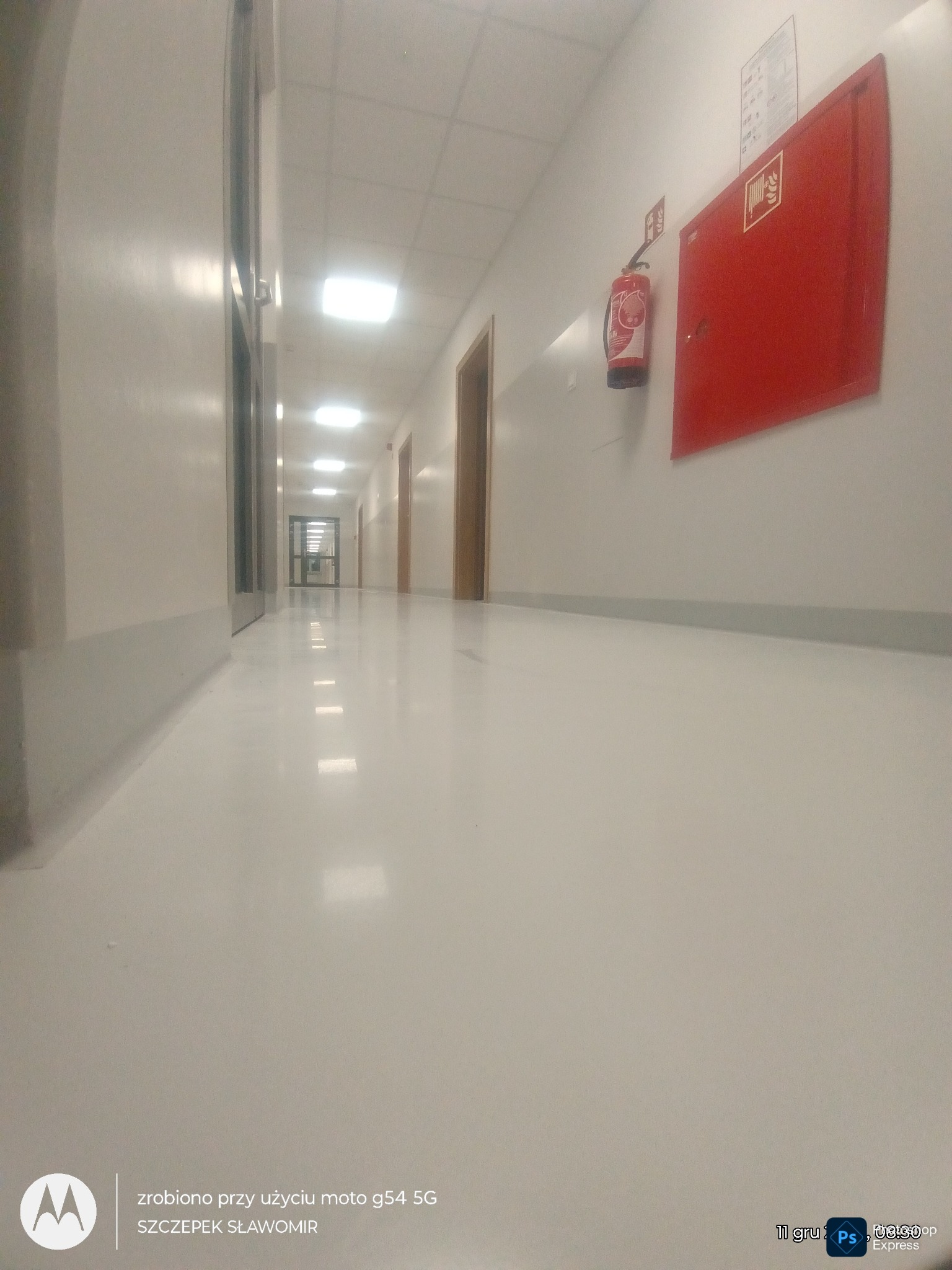 Długi, jasny korytarz z białymi ścianami i podłogą. Na ścianie widoczna czerwona szafka hydrantowa i gaśnica. Perspektywa z niskiego kąta, widoczne drzwi na końcu korytarza.