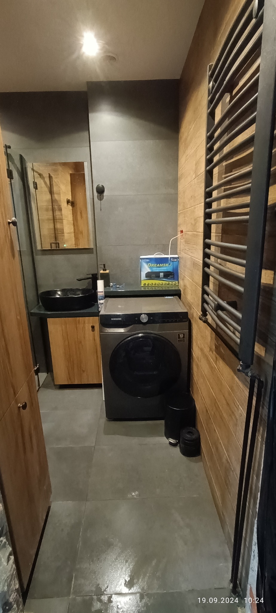 Szara łazienka z drewnianymi akcentami: umywalka na szafce, pralka Samsung, grzejnik drabinkowy i szare płytki podłogowe. Na pralce pudełko DreamSky.