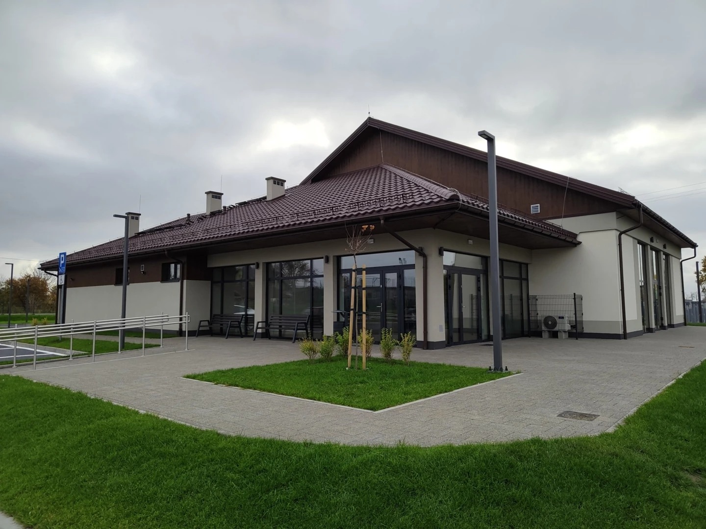 Nowoczesny budynek Centrum Kultury w Wielkiej Łące z brązowym dachem, dużymi oknami i zielonym trawnikiem. Widoczny podjazd dla niepełnosprawnych i parking.