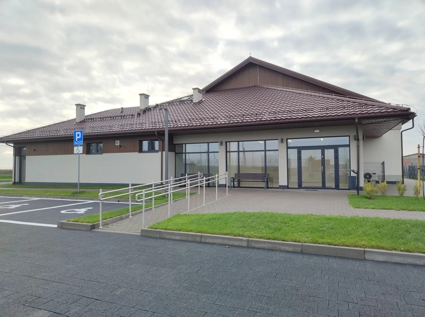 Nowoczesny budynek centrum kultury z brązowym dachem, rampą dla niepełnosprawnych i parkingiem. Widoczne duże okna i ławka przed wejściem. Pochmurne niebo w tle.