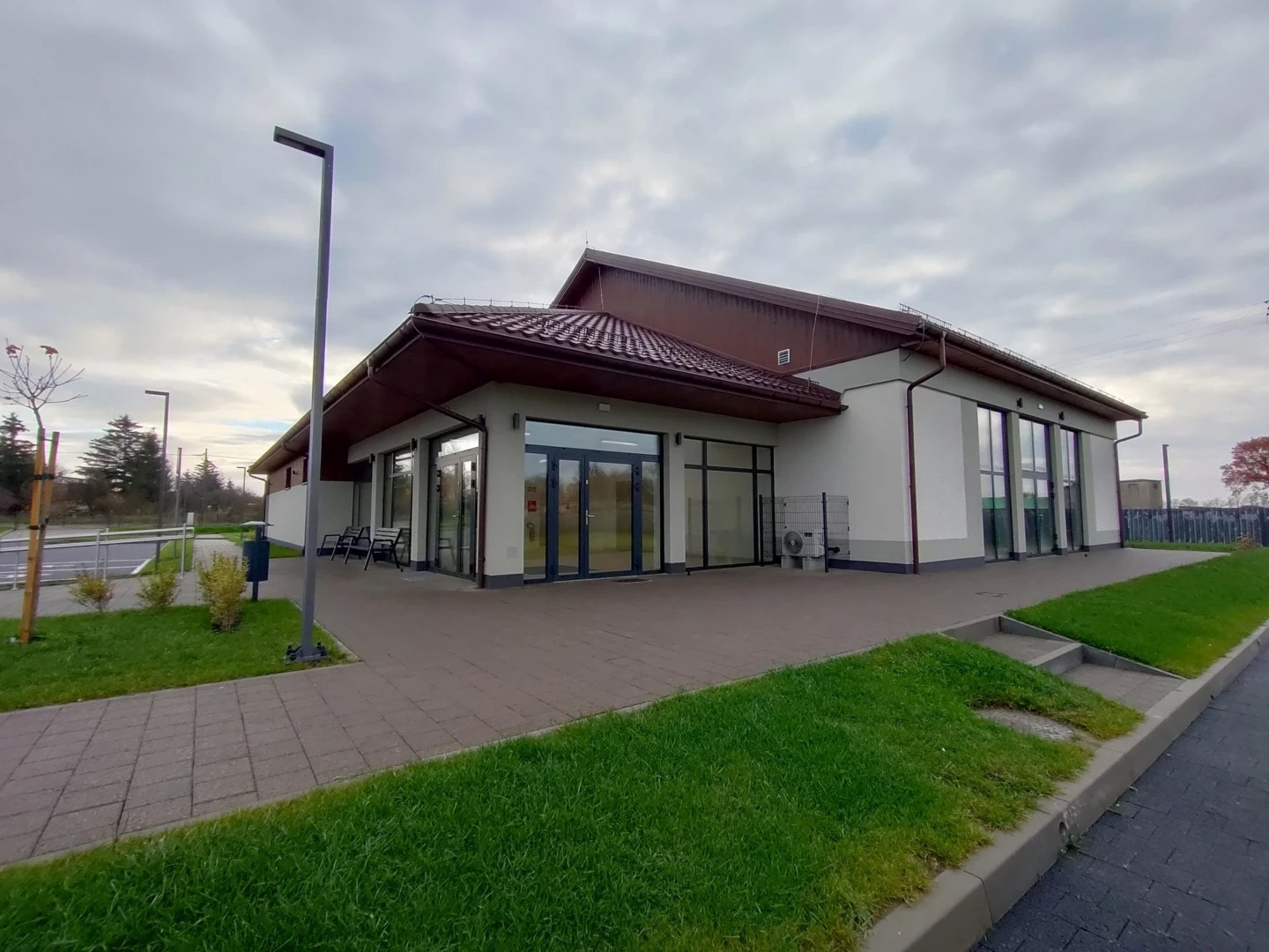 Nowoczesny budynek centrum kultury z brązowym dachem i dużymi oknami, otoczony zielenią i chodnikiem z kostki. Widok z zewnątrz w pochmurny dzień.