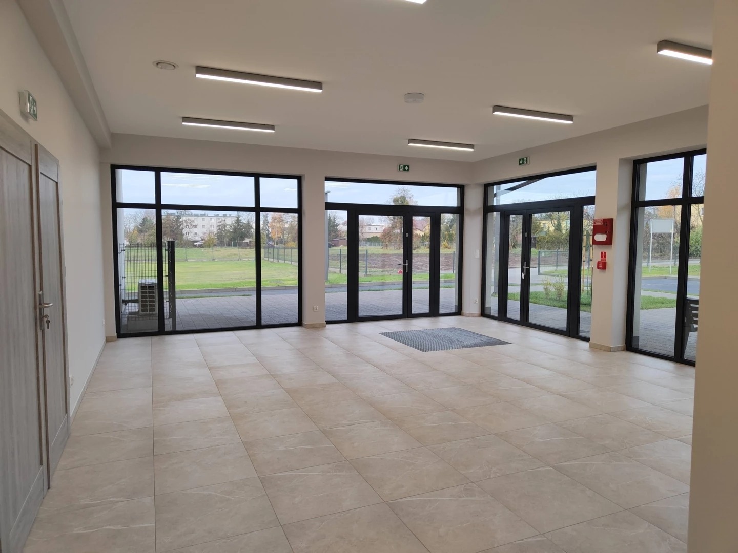 Jasne, przestronne wnętrze z beżową podłogą, dużymi oknami z czarnymi ramami i nowoczesnym oświetleniem sufitowym. Widok na zewnątrz z zielenią i budynkami w tle.