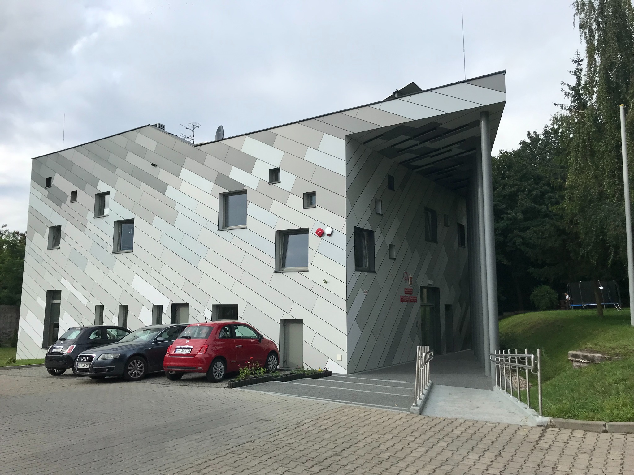 Nowoczesny budynek szkoły specjalnej z elewacją w odcieniach szarości, z trzema zaparkowanymi samochodami i zielenią w tle. Uwagę zwraca geometryczny design elewacji.