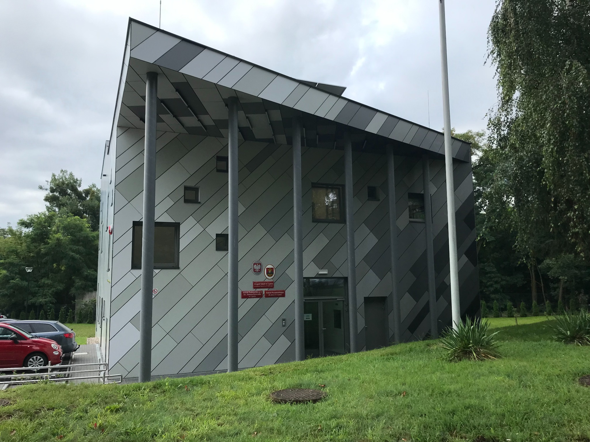 Szkoła Specjalna w Lipnie: nowoczesny budynek z szarej, ukośnej elewacji, wsparty na kolumnach. Widoczne logo szkoły i flaga. Zieleń trawnika i drzewa w tle.