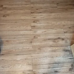 Jasne, drewnopodobne panele podłogowe ułożone w regularny wzór, z widoczną fakturą drewna i delikatnymi przebarwieniami. Ujęcie z góry, fragment podłogi.