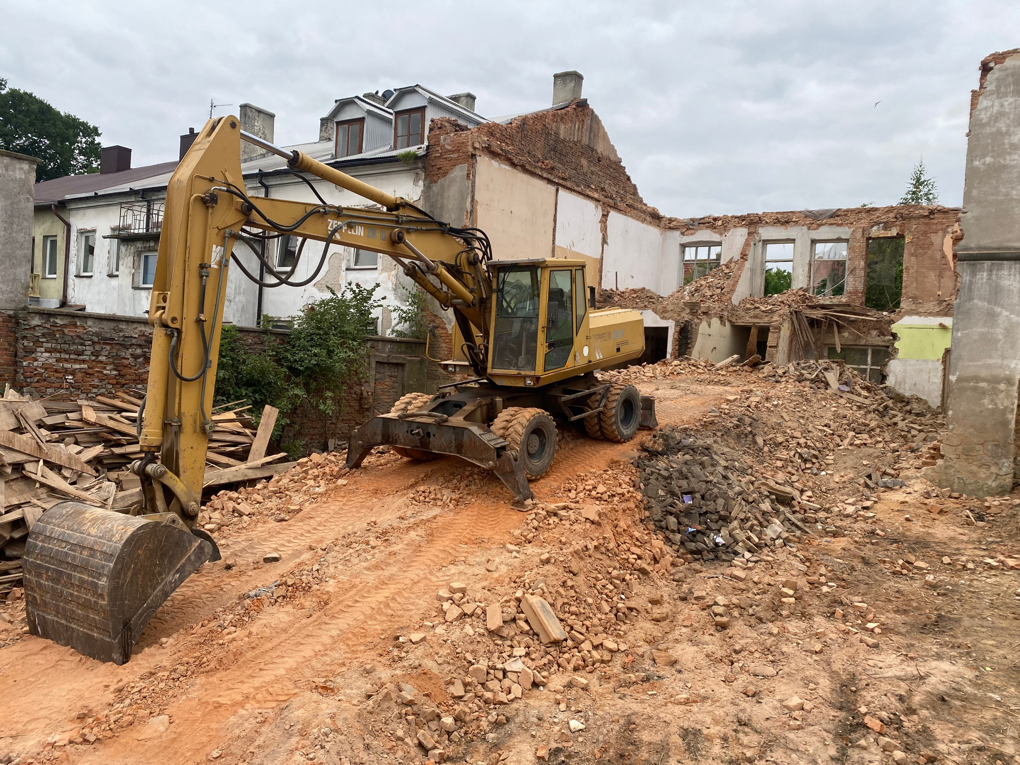 Żółta koparka kołowa marki Zeppelin na tle częściowo rozebranego, ceglanego budynku. Widoczne gruz i drewniane elementy.