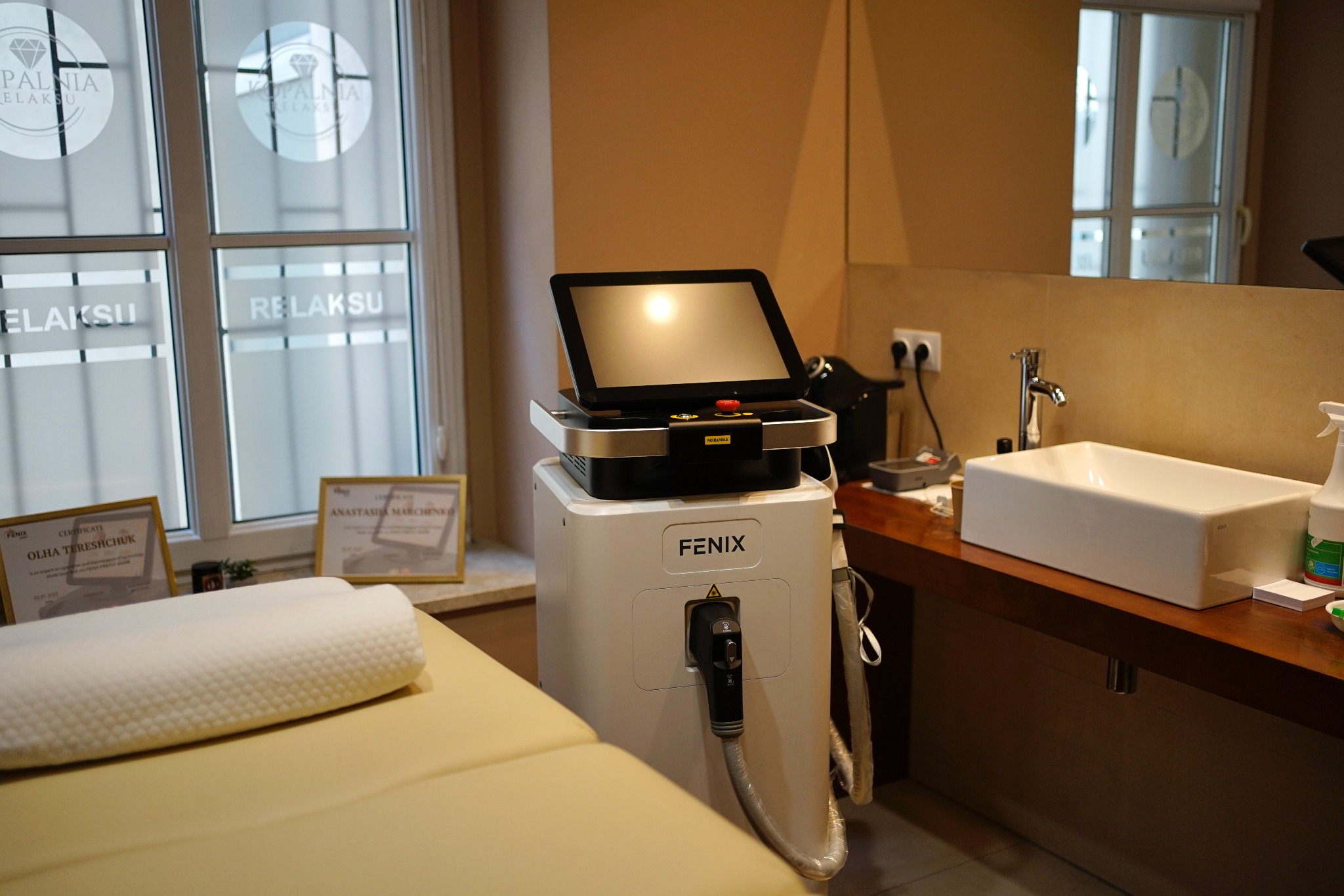 Wnętrze gabinetu kosmetycznego: łóżko zabiegowe, urządzenie Fenix, okno z logo, umywalka i certyfikaty w ramkach. Przytulna aranżacja.