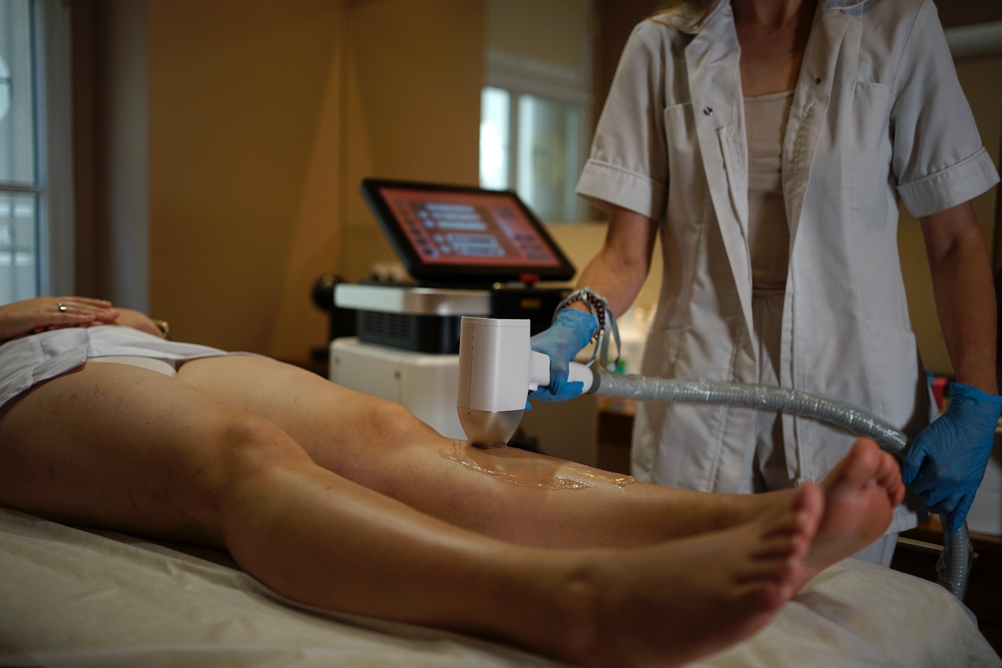 Zabieg z użyciem lasera na nodze pacjentki leżącej na leżance. Kosmetolog w rękawicach ochronnych, w tle widoczny ekran urządzenia i okno.