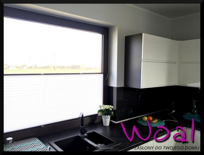 Jasna plisa okienna w nowoczesnej kuchni z czarnym blatem i białymi szafkami. Widok z okna na zewnątrz. Minimalistyczny styl.