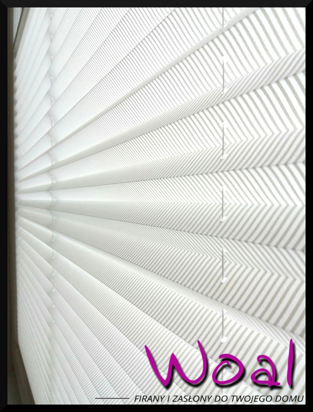Biała plisa okienna z delikatnym szarym wzorem w paski, zamontowana w oknie. Widoczne detale konstrukcji i fałdy materiału. U dołu logo firmy.