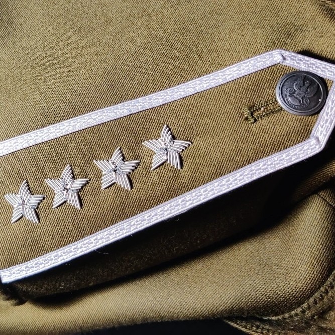 Zbliżenie na naramiennik munduru wojskowego w odcieniach khaki, z czterema srebrnymi gwiazdkami i guzikiem z orłem, ukazujący precyzyjne haftowanie stopnia.