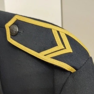 Fragment munduru PSP z naszywką stopnia na ramieniu, widoczny guzik. Detal obszycia w kolorze złotym na ciemnym tle tkaniny mundurowej.