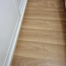 Parkiet z jasnego drewna ułożony przy białej ścianie i listwie przypodłogowej. Widoczna struktura drewna i delikatne łączenia desek. Minimalistyczne wykończenie podłogi.