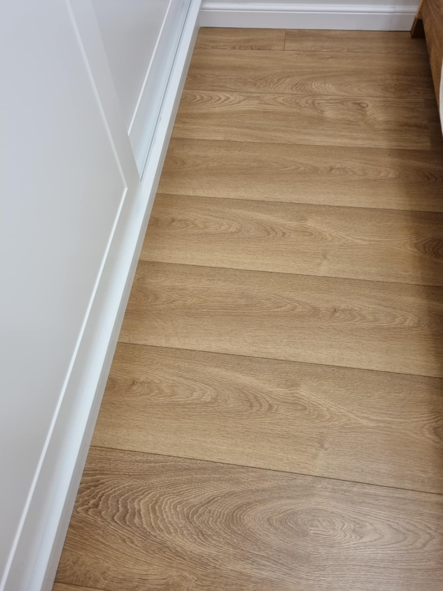 Parkiet z jasnego drewna ułożony przy białej ścianie i listwie przypodłogowej. Widoczna struktura drewna i delikatne łączenia desek. Minimalistyczne wykończenie podłogi.