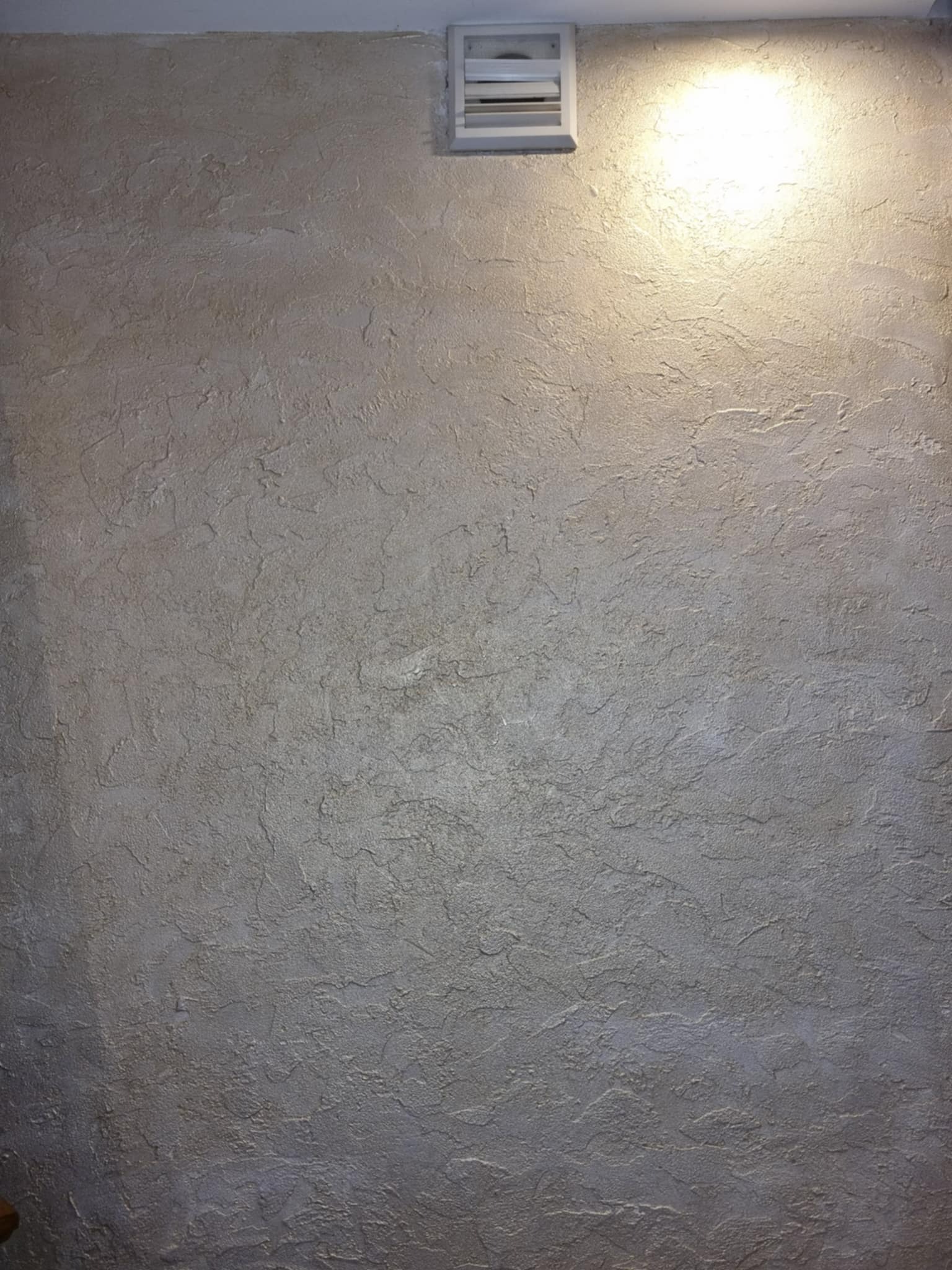 Beżowa ściana z fakturą, oświetlona od góry, z kratką wentylacyjną w rogu. Tekstura ściany przypomina naturalny kamień.