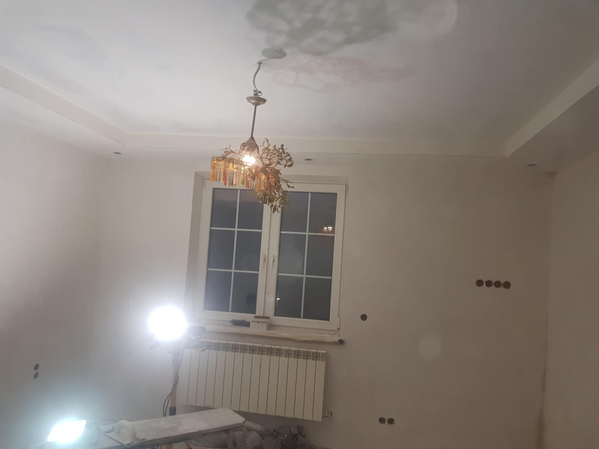 Wykończone ściany i sufit w pokoju z oknem, widoczny ozdobny żyrandol, grzejnik i przygotowane punkty elektryczne. Światło robocze oświetla wnętrze.