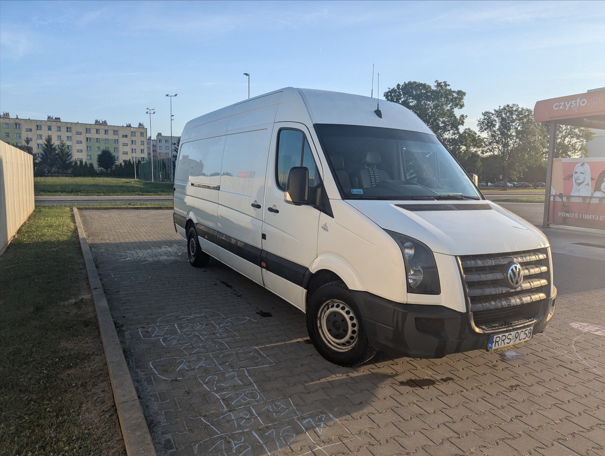 Biały bus dostawczy Volkswagen Crafter zaparkowany na kostce brukowej z numerem rejestracyjnym RRS 9C58, w tle budynek mieszkalny i myjnia.