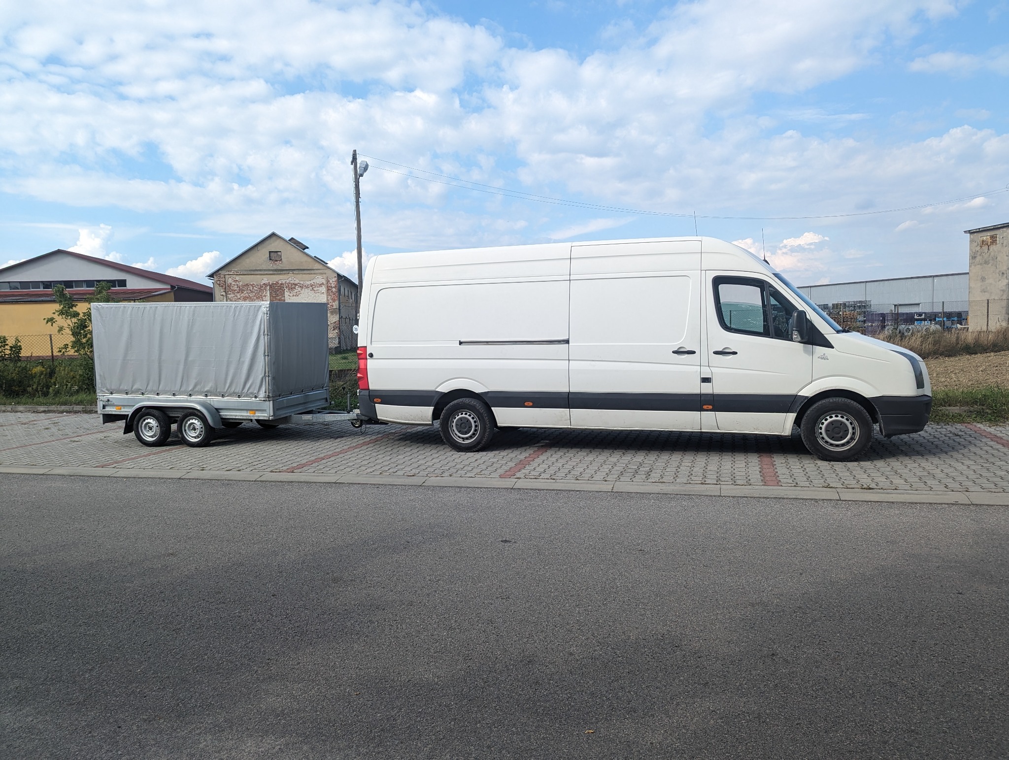 Biały van z przyczepą krytą plandeką na parkingu. Przyczepa ma dwa koła. W tle budynki i błękitne niebo z chmurami. Usługa transportowa z wykorzystaniem przyczepy.