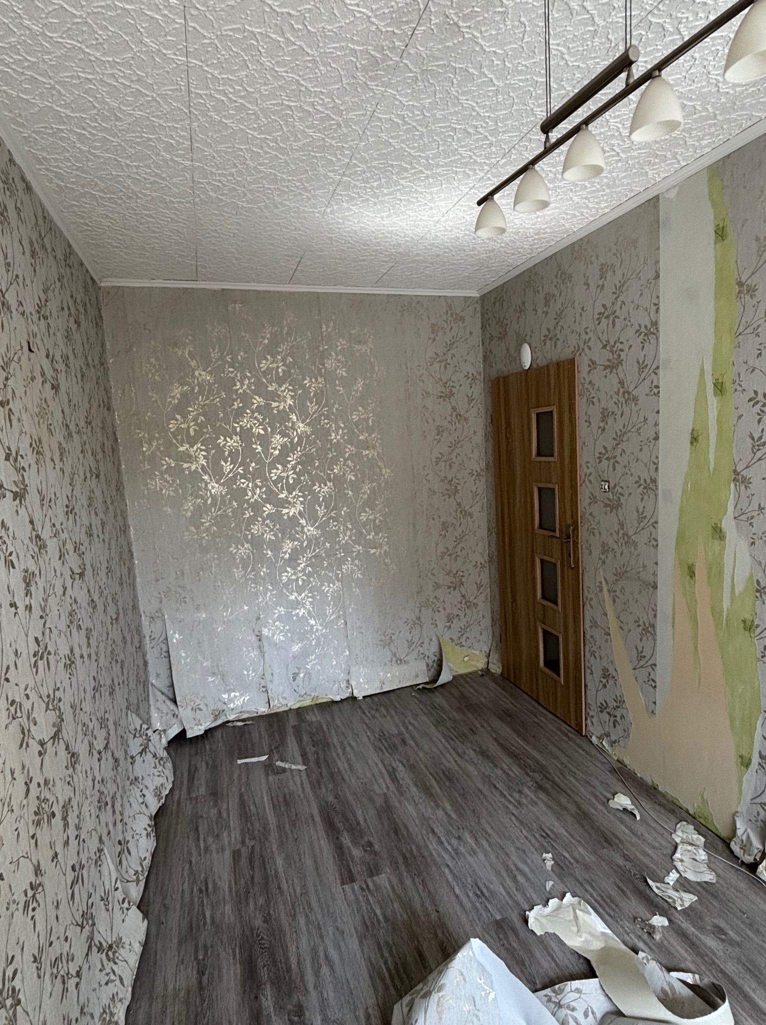 Pokój przed remontem: zerwane tapety z kwiatowym wzorem na ścianach i fragmenty na podłodze, widoczny sufit z fakturą i lampa sufitowa. Przygotowanie do malowania.