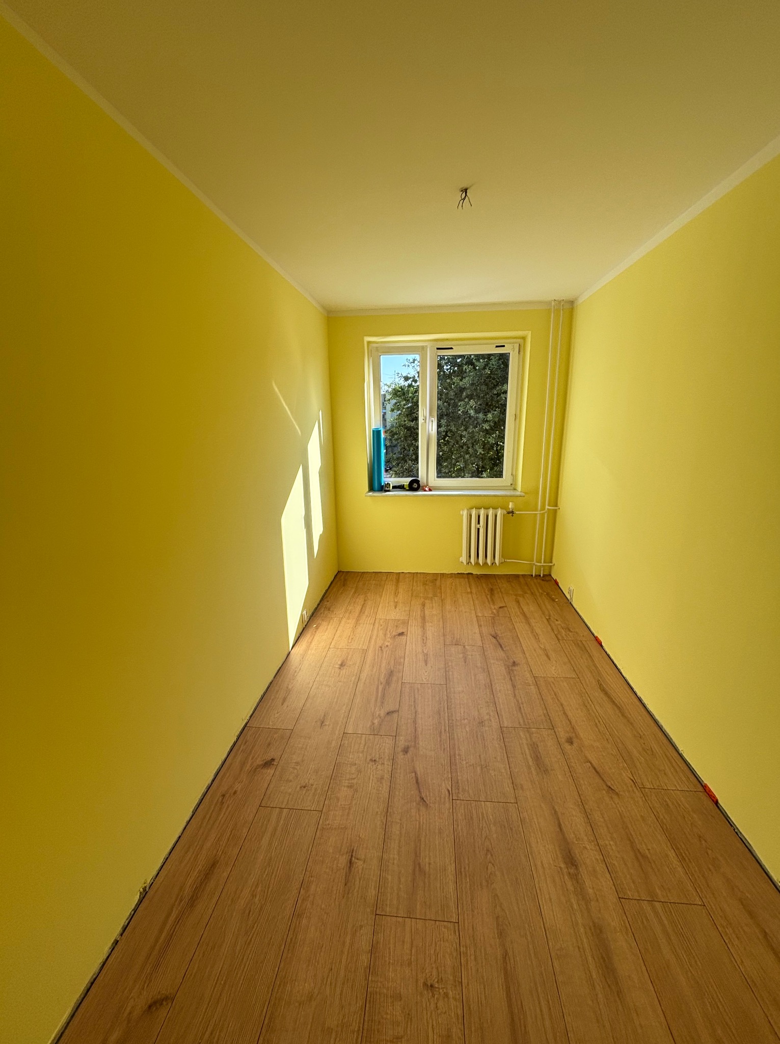 Pomalowane na żółto wnętrze pokoju z drewnianą podłogą i oknem z widokiem na drzewa. Świeżo wykończone ściany, widoczny kaloryfer pod oknem.