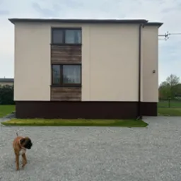 Dwukondygnacyjny dom z beżową elewacją, brązowym cokołem i drewnianą wstawką między oknami. Na pierwszym planie pies rasy bokser na żwirowym podłożu.