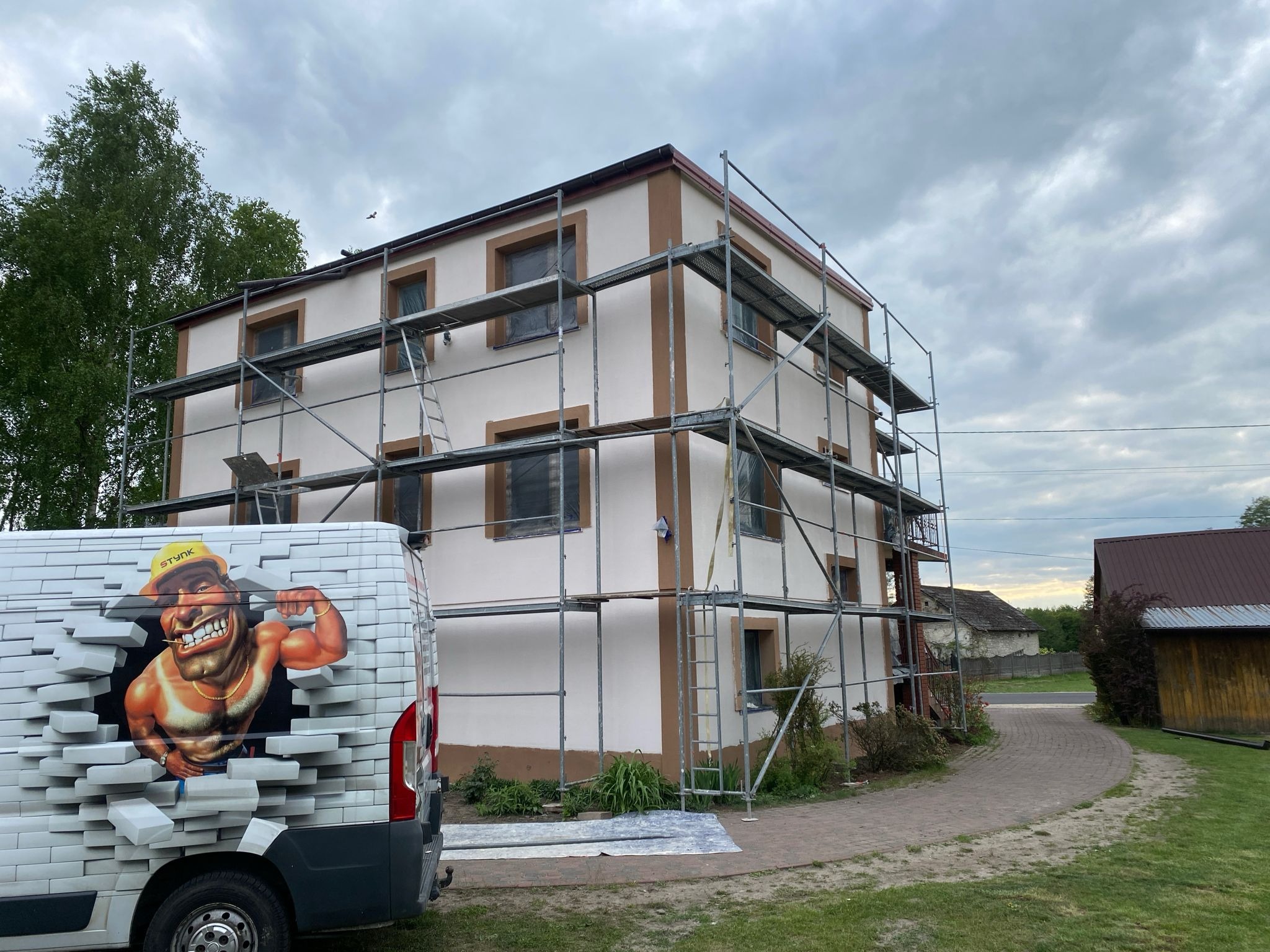 Dwukondygnacyjny dom w rusztowaniach, częściowo pomalowany na biało z brązowymi obramowaniami okien, obok zaparkowany samochód z grafiką silnego murarza.
