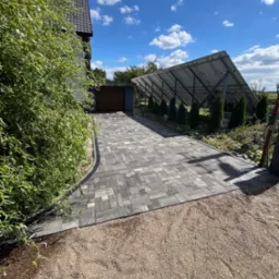 Ułożona, szara kostka brukowa prowadząca do garażu pod panele słoneczne. Krawężnik oddziela kostkę od żwirowego podłoża. W tle błękitne niebo z chmurami.