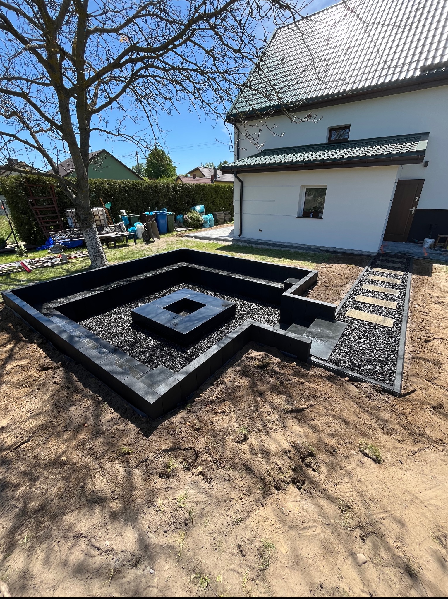 Nowoczesna aranżacja ogrodu z czarną kostką i paleniskiem, widok z góry. Minimalistyczny design, schludne wykonanie, jasny dom w tle.